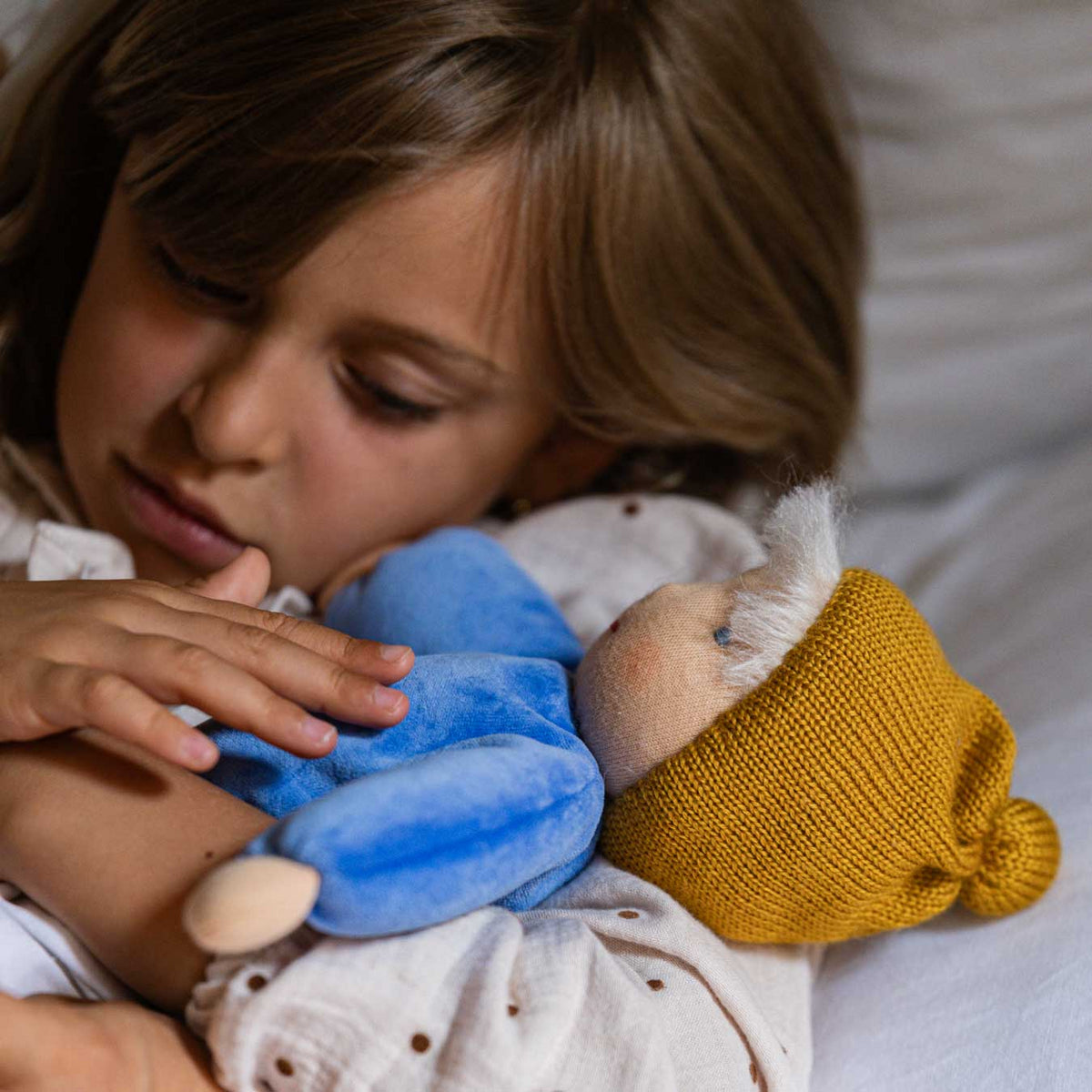 Kind kuschelt mit der Käthe Kruse Waldorfpuppe NANA wolke mit blauem Körper und senfgelber Strickmütze