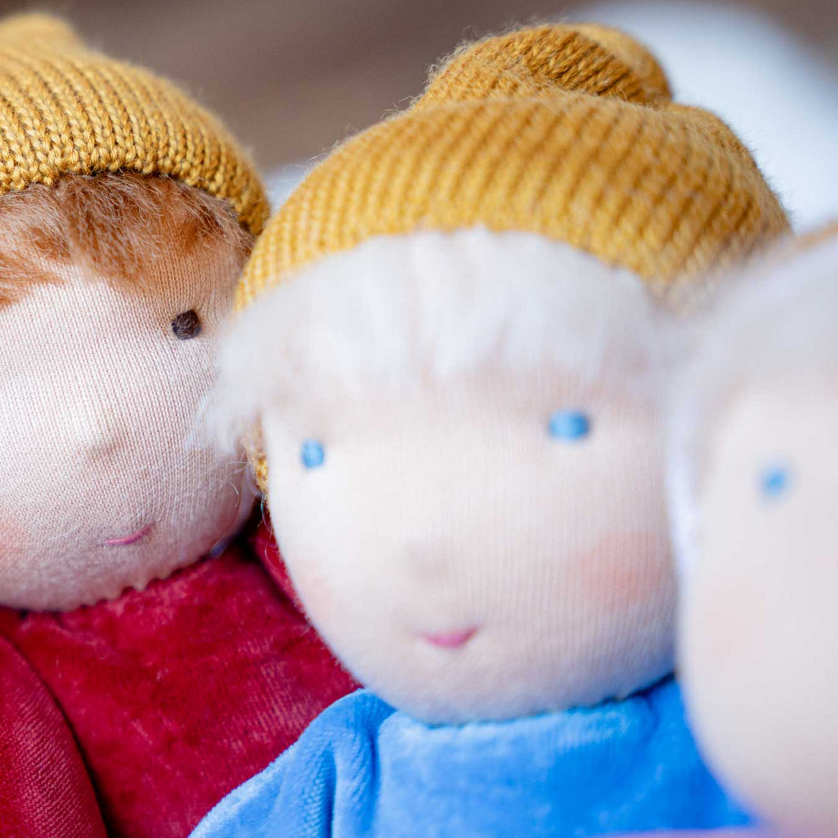 Nahaufnahme der Käthe Kruse Waldorfpuppe NANA wolke mit hellem Haar und senfgelber Mütze zwischen zwei weiteren Nanas