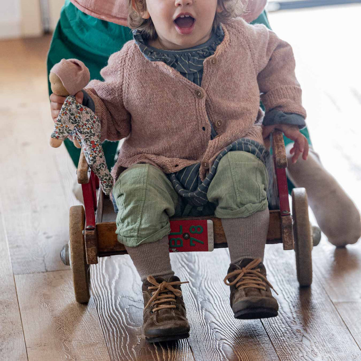 Kleinkind sitzt in einem Holzwagen und hält die Käthe Kruse Waldorfpuppe NINI Blümchen in der Hand