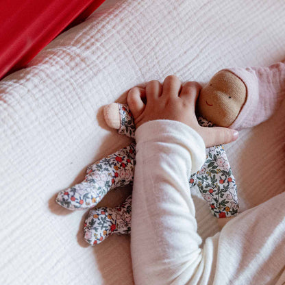 Kinderhände halten die Käthe Kruse Waldorfpuppe NINI Blümchen im geblümten Anzug auf einer weißen Decke