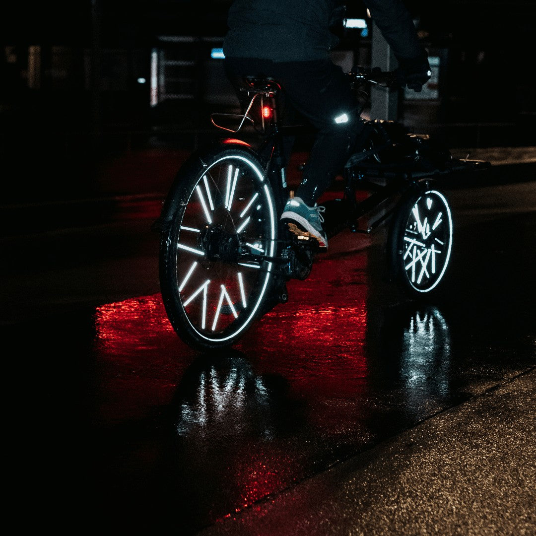 Fahrrad bei Nacht, die großen Rainette Speichenreflektoren leuchten weiß, darunter roter Lichtschein auf nassem Boden