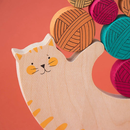Londji Meow Holzkatze beige mit gelben Ohren und Streifen neben bunten Wollknäueln auf Korallenhintergrund